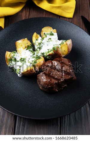 Grilled meat steak with baked potatoes, cream sauce and fresh herbs. Dish on a wooden dark background with a bright yellow napkin.