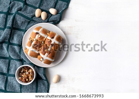 Delicious vegan homemade hot cross buns on a plate. The plate sits on a navy coloured tea towel and is surrounded by a bowl of various nuts and wooden easter eggs. Celebrate the Easter Holidays.