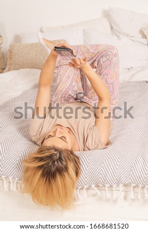 A girl in pajamas is lying on a bed in her apartment with a phone. She is surfing in social networks and smiling. Soft pink creamy colors of the picture. Vertical shot