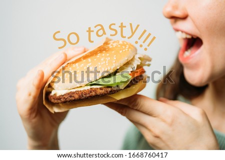 The girl holds a hamburger with her hands and eats it with appetite. The inscription is so tasty. Close up.