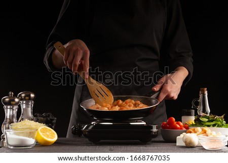 Chef fries seafood, shrimp in a pan. Against the background of vegetables. Black background, cooking and recipe book, cooking and healthy eating.