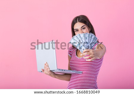Image of cheerful young woman standing isolated over pink background using laptop computer and holding money banknotes . Portrait of a smiling girl holding laptop computer