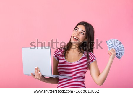 Portrait of an excited satisfied girl holding money banknotes and laptop computer. Happy woman with lucky money. Photo of beautiful woman with laptop, isolated on pink background