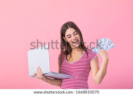 Image of cheerful young woman standing isolated over pink background using laptop computer and holding money banknotes . Portrait of a smiling girl holding laptop computer
