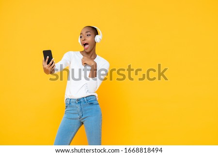 Confident African American woman wearing headphones listening to music from mobile phone in yellow isolated studio background