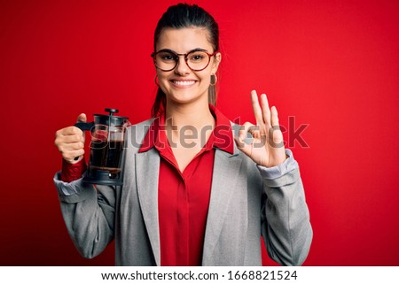 Young beautiful brunette woman doing coffe holding french coffeemaker over red background doing ok sign with fingers, excellent symbol