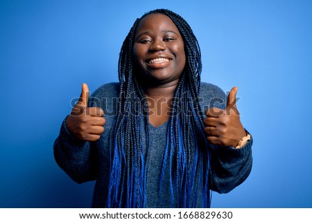 African american plus size woman with braids wearing casual sweater over blue background success sign doing positive gesture with hand, thumbs up smiling and happy. Cheerful expression and winner 