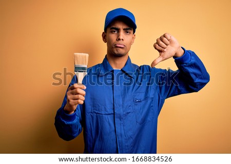 Young handsome african american painter man wearing uniform using painting brush with angry face, negative sign showing dislike with thumbs down, rejection concept
