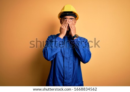 Young handsome african american worker man wearing blue uniform and security helmet rubbing eyes for fatigue and headache, sleepy and tired expression. Vision problem