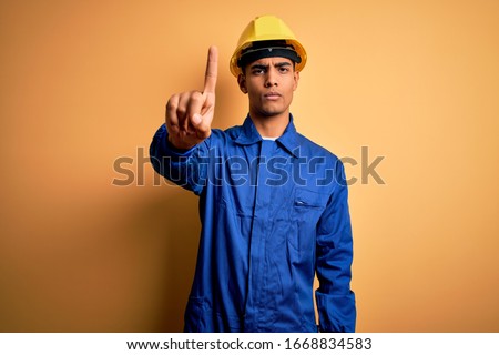 Young handsome african american worker man wearing blue uniform and security helmet Pointing with finger up and angry expression, showing no gesture