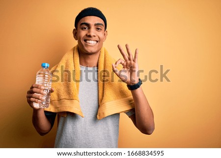 Young handsome african american sportsman wearing towel drinking bottle of water doing ok sign with fingers, excellent symbol