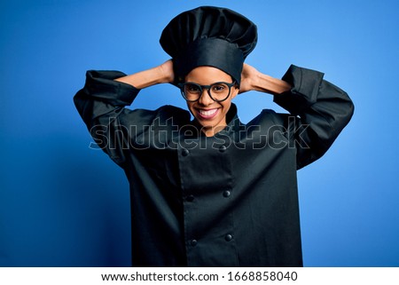 Young african american chef woman wearing cooker uniform and hat over blue background relaxing and stretching, arms and hands behind head and neck smiling happy