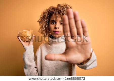 Young african american curly woman holding bowl with chips potatoes over yellow background with open hand doing stop sign with serious and confident expression, defense gesture