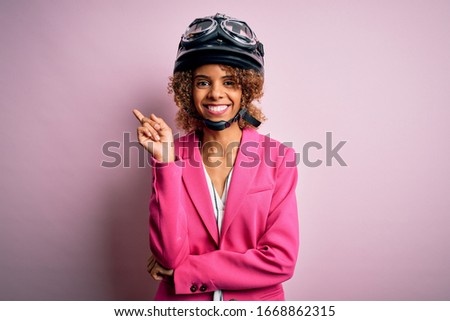 African american motorcyclist woman with curly hair wearing moto helmet over pink background with a big smile on face, pointing with hand and finger to the side looking at the camera.