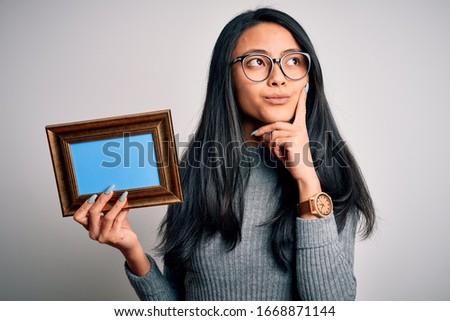 Young beautiful chinese woman holding vintage frame over isolated white background serious face thinking about question, very confused idea