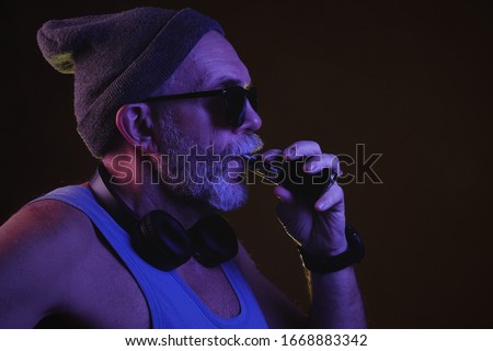 Side view stock footage of a hipster aged man in grey hat, blue tank top, sunglasses and headphones around neck vaping an electronic cigarette. Isolate on dark background.