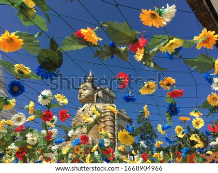Large shining golden buddha in focus in the background with a soft off-focus on the red, yellow, white and blue flower hung net in front with clear blue skies