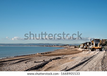 Landscape shot of pebble beach on a sunny day with calm blue sea and land on the horizon.  Large lorries to the right of the picture coming towards view.