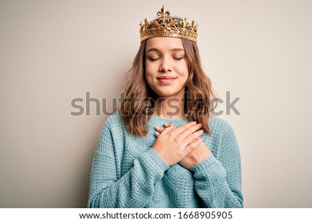 Young blonde girl wearing queen golden crown over isolated background smiling with hands on chest with closed eyes and grateful gesture on face. Health concept.