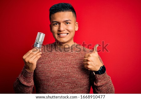 Young handsome latin man holding led lightbulb over isolated red background happy with big smile doing ok sign, thumb up with fingers, excellent sign