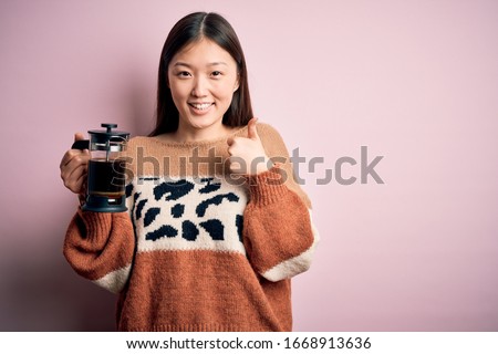 Young asian woman making a glass of coffe using french press coffee maker over pink background happy with big smile doing ok sign, thumb up with fingers, excellent sign