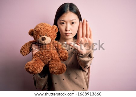 Young asian woman hugging teddy bear stuffed animal over pink isolated background with open hand doing stop sign with serious and confident expression, defense gesture