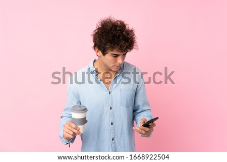 Young caucasian man over isolated pink background holding coffee to take away and a mobile