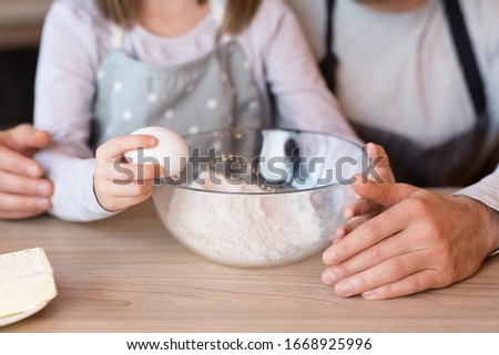 Father and daughter preparing dough together in kitchen, adding eggs to bowl while cooking pastry at home, cropped image, closeup