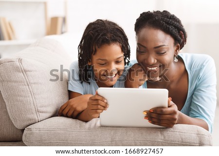African Mom And Daughter Watching Cartoons Together On Digital Tablet, Lying On Couch. Free space