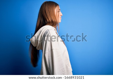 Young beautiful redhead sporty woman wearing sweatshirt over isolated blue background looking to side, relax profile pose with natural face with confident smile.