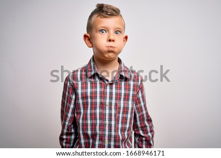 Young little caucasian kid with blue eyes wearing elegant shirt standing over isolated background puffing cheeks with funny face. Mouth inflated with air, crazy expression.