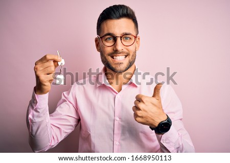 Young real estate business man holding new house keys over pink background happy with big smile doing ok sign, thumb up with fingers, excellent sign
