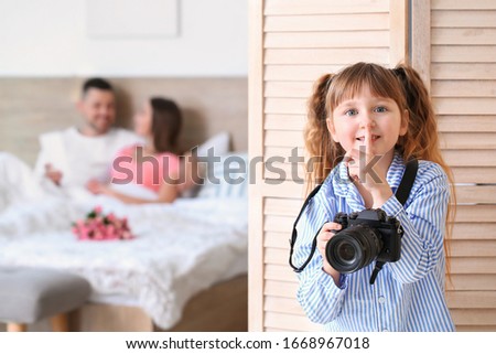 Little girl with photo camera hiding behind folding screen in bedroom of her parents