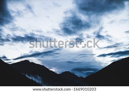 Low cloud above mountain lake. Mysterious silhouettes of big mountains and trees. Dense fog above firs and pines. Highland tranquil landscape at early morning. Ghostly alpine atmospheric scenery.