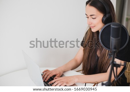 Attractive young woman blogger with long black hair recording online podcast using her laptop, headphones and professional microphone. Place for text.