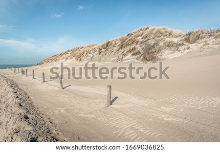 Sand path through the dunes to the Dutch North Sea beach. The path is covered with drifting sand. Marram grass grows on the dunes. Along the path is a fence of wooden posts and barbed wire.