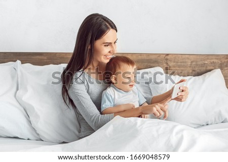 Motherhood. Mother and son at home sitting in bed taking selfie on smartphone smiling joyful