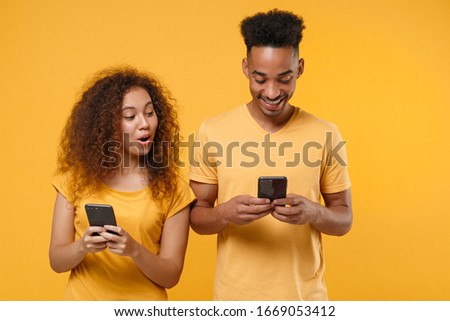 Amazed young friends couple african american guy girl in casual clothes isolated on yellow orange wall background. People lifestyle concept. Mock up copy space. Using mobile phone, typing sms message