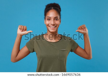 Smiling young african american woman girl in casual t-shirt posing isolated on blue background studio portrait. People sincere emotions lifestyle concept. Mock up copy space. Point thumbs on herself