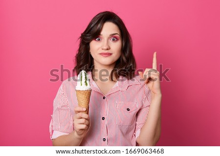 Indoor portrait of magnetic joyful charming black haired female standing isolated over pink background in studio, making gesture, raising one finger, having idea, tasting ice cream. Food concept.