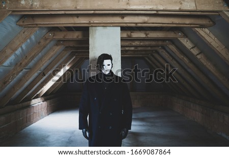 A scary guy in white mask with black hair and long dark coat standing in an abandoned attic. Scary and mystical picture. Horror concept.