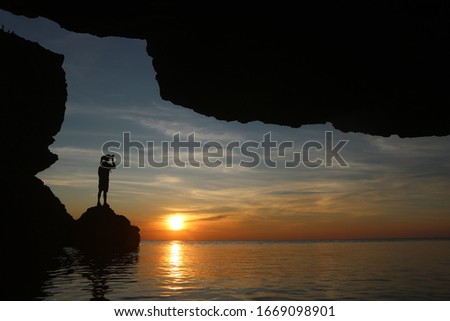 Silhouette of photographer using the camera to capture the sunset in a cave by the sea at twilight sky sunset.
