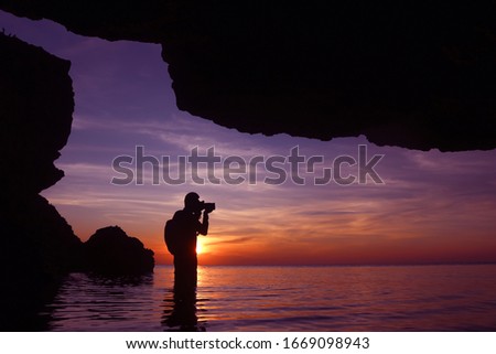Silhouette of photographer using the camera to capture the sunset in a cave by the sea at twilight sky sunset.
