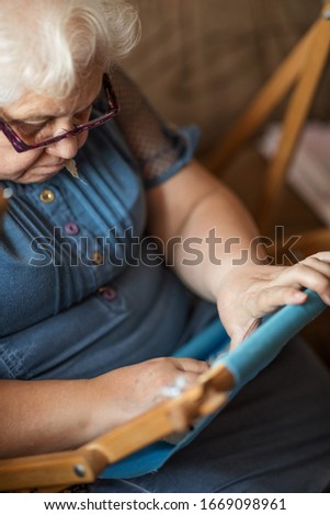Selective focus. A woman of 74 years old is engaged in embroidery on an embroidery frame in a blue dress sitting on a sofa, in daylight. Great picture of the concept of Hobbies and Hobbies at any age.