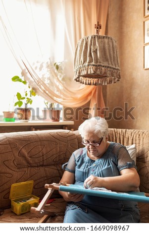 Selective focus. A woman of 74 years old is engaged in embroidery on an embroidery frame in a blue dress sitting on a sofa, in daylight. Great picture of the concept of Hobbies and Hobbies at any age.