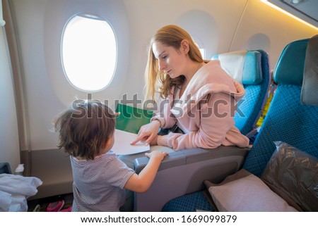  young beautiful mother sits in an airplane chair shows with finger a page in a book to her little cute toddler, who is standing in front of her. close-up, soft focus, top view. copy space,blank page