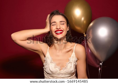 Surprised young attractive dark haired female with evening makeup smiling widely with closed eyes and holding raised hand on her head, rejoicing while celebrating something over burgundy background