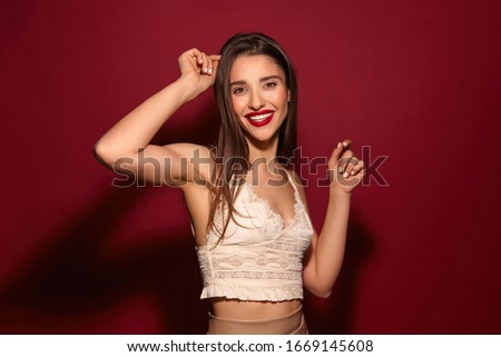 Cheerful young attractive long haired brunette female with evening makeup raising her hands while dancing and smiling happily at camera, isolated over burgundy background