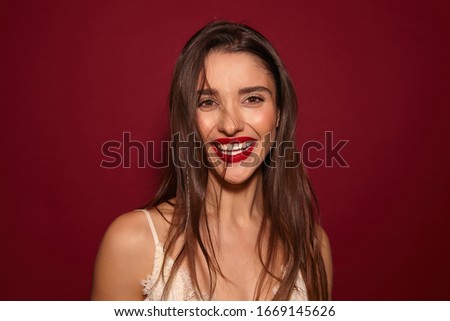 Indoor photo of young attractive dark haired female with evening makeup and wild hairstyle looking cheerfully at camera with broad smile, posing over burgundy background