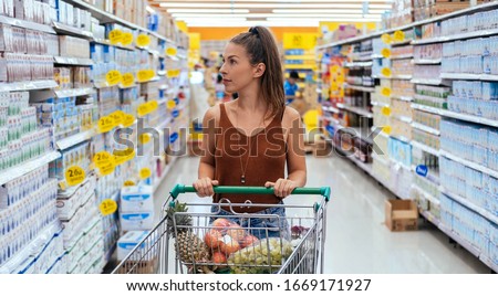 Smiling woman pushing a full shopping cart at supermarket stock photo. Beautiful young woman shopping healthy food in a grocery store/supermarket. Retail, sale and consumerism concept